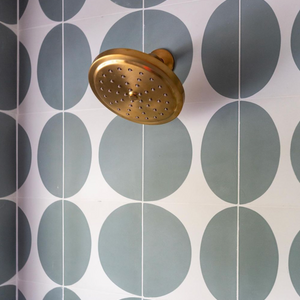 A brass showerhead on a tiled wall with Pomelo Zeppelin 8x8 tiles—bold green circles on white—brings a playful, midcentury modern vibe to the space.