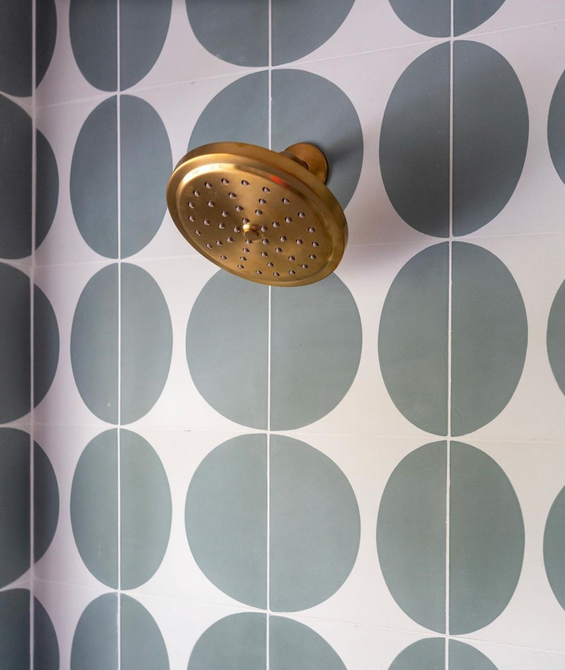A brass showerhead on a tiled wall with Pomelo Zeppelin 8x8 tiles—bold green circles on white—brings a playful, midcentury modern vibe to the space.
