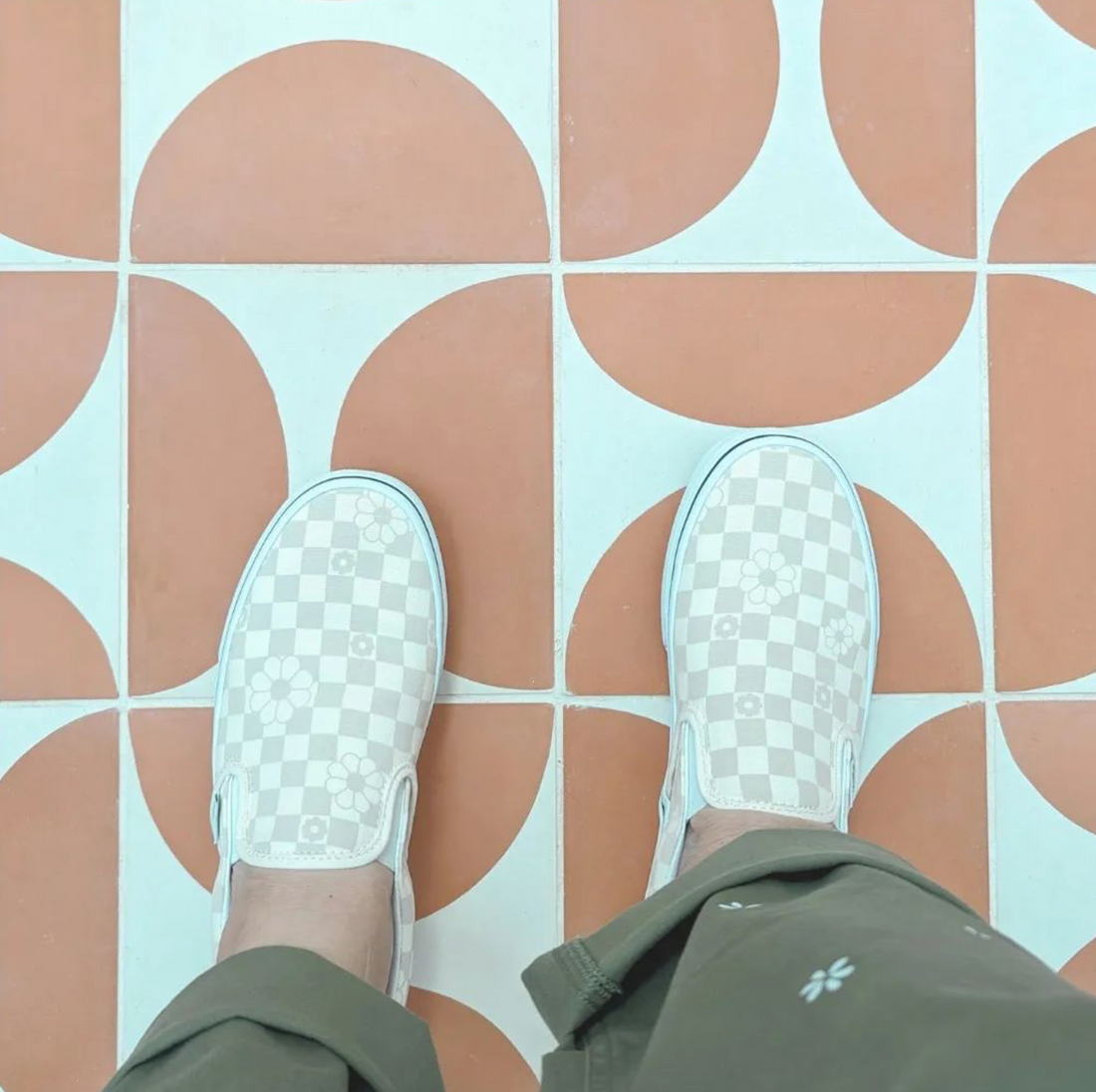 A person in olive green pants and light-colored checkered slip-ons stands on Pomelo Jaipur Pink 8x8 tile featuring a geometric orange and white circular pattern.
