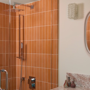 A modern bathroom with Terra Cotta 2x8 tiles in the shower, a glass door, wall-mounted showerhead, marble countertop, decorative vases, round mirror, and a textured glass wall sconce.