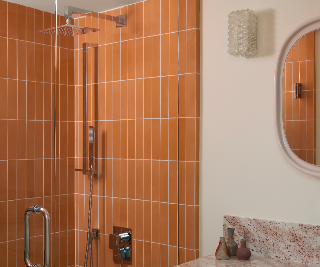A modern bathroom with Terra Cotta 2x8 tiles in the shower, a glass door, wall-mounted showerhead, marble countertop, decorative vases, round mirror, and a textured glass wall sconce.