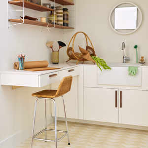A tidy craft room with a white counter, farmhouse sink, Press Play Bone 4x8 tile backsplash, wicker chair, and wall shelves with jars and baskets. Art supplies, vegetables, and gardening gloves sit under a round mirror.