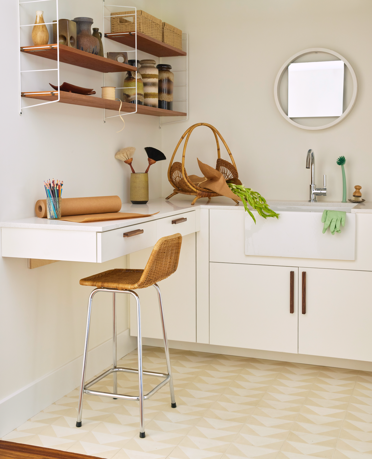 A tidy craft room with a white counter, farmhouse sink, Press Play Bone 4x8 tile backsplash, wicker chair, and wall shelves with jars and baskets. Art supplies, vegetables, and gardening gloves sit under a round mirror.