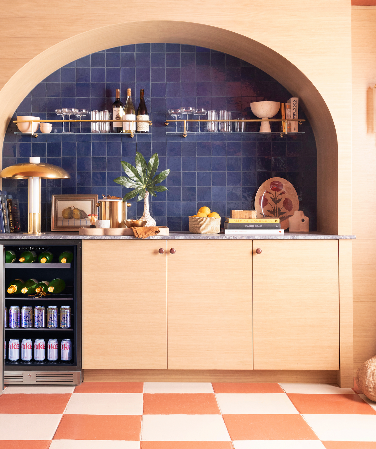 A stylish home bar with light wood cabinets, Night Blue 4x4 tiles by vendor-unknown as a backsplash, gold accents, wine bottles, glassware, a mini fridge with drinks, and decor items beneath a large arched alcove.