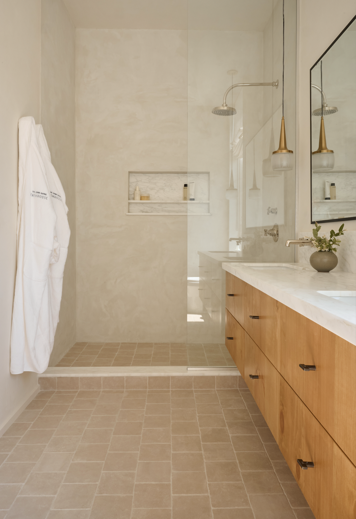 A modern bathroom features Zia Tile’s Buff 6x6 + Honed beige floor tiles, a glass shower, floating wood vanity with white countertop, large mirror, pendant lights, and a white robe hanging on the wall.