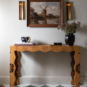 A wooden console table stands against a light wall with books, a cup, and a dark vase. Above, a framed windmill painting and two modern sconces echo the Stone 8x8 cement tile backsplash style.