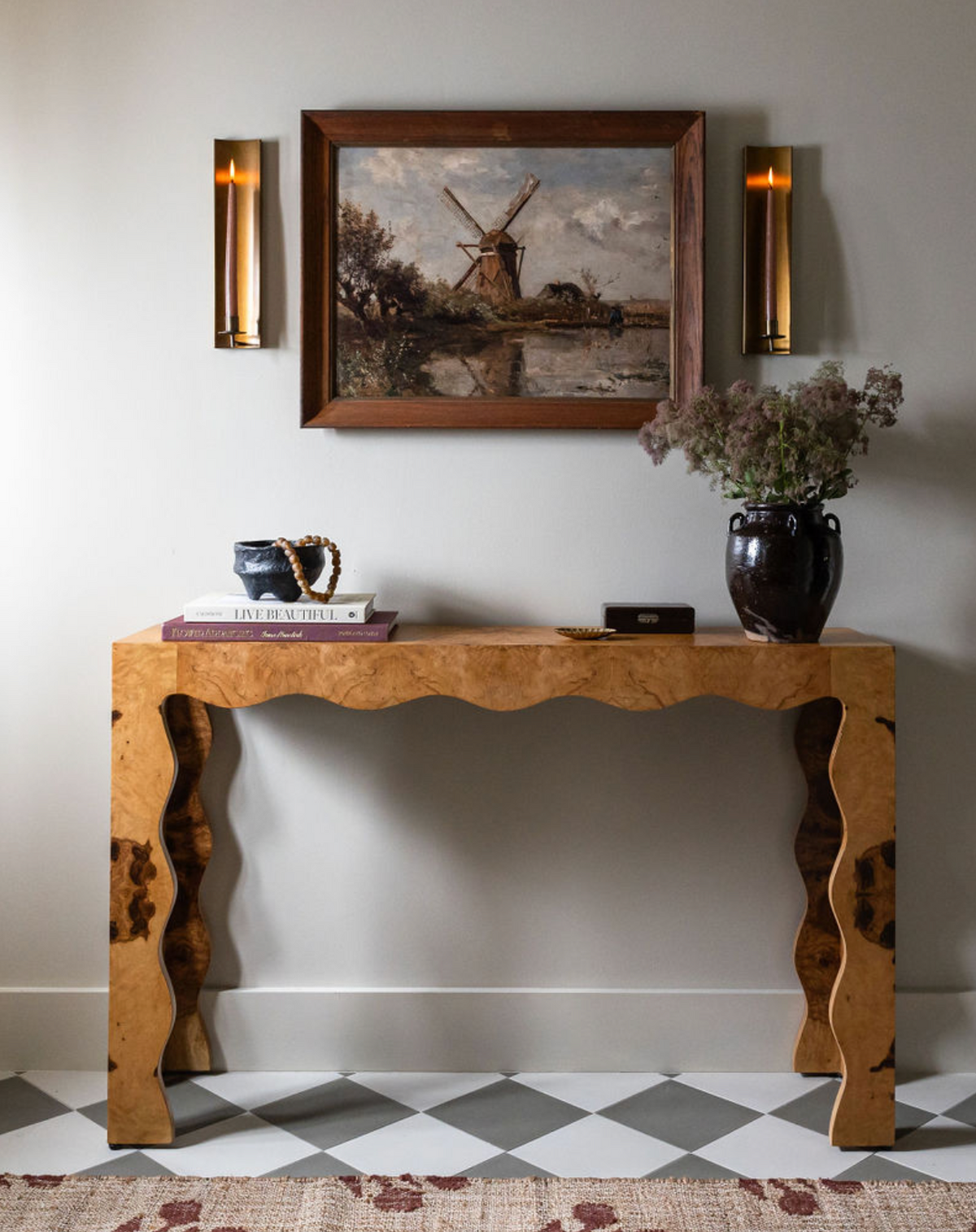 A wooden console table stands against a light wall with books, a cup, and a dark vase. Above, a framed windmill painting and two modern sconces echo the Stone 8x8 cement tile backsplash style.