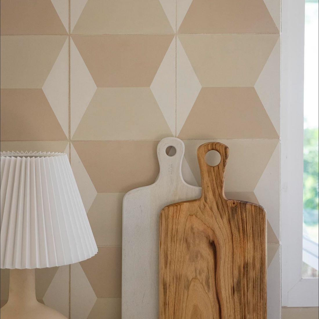 Two wooden cutting boards, one white and one natural wood, lean against a wall with Zia Tiles Colorama Bone 8x8 tiles, beside a cream table lamp with a pleated shade on a marble countertop.