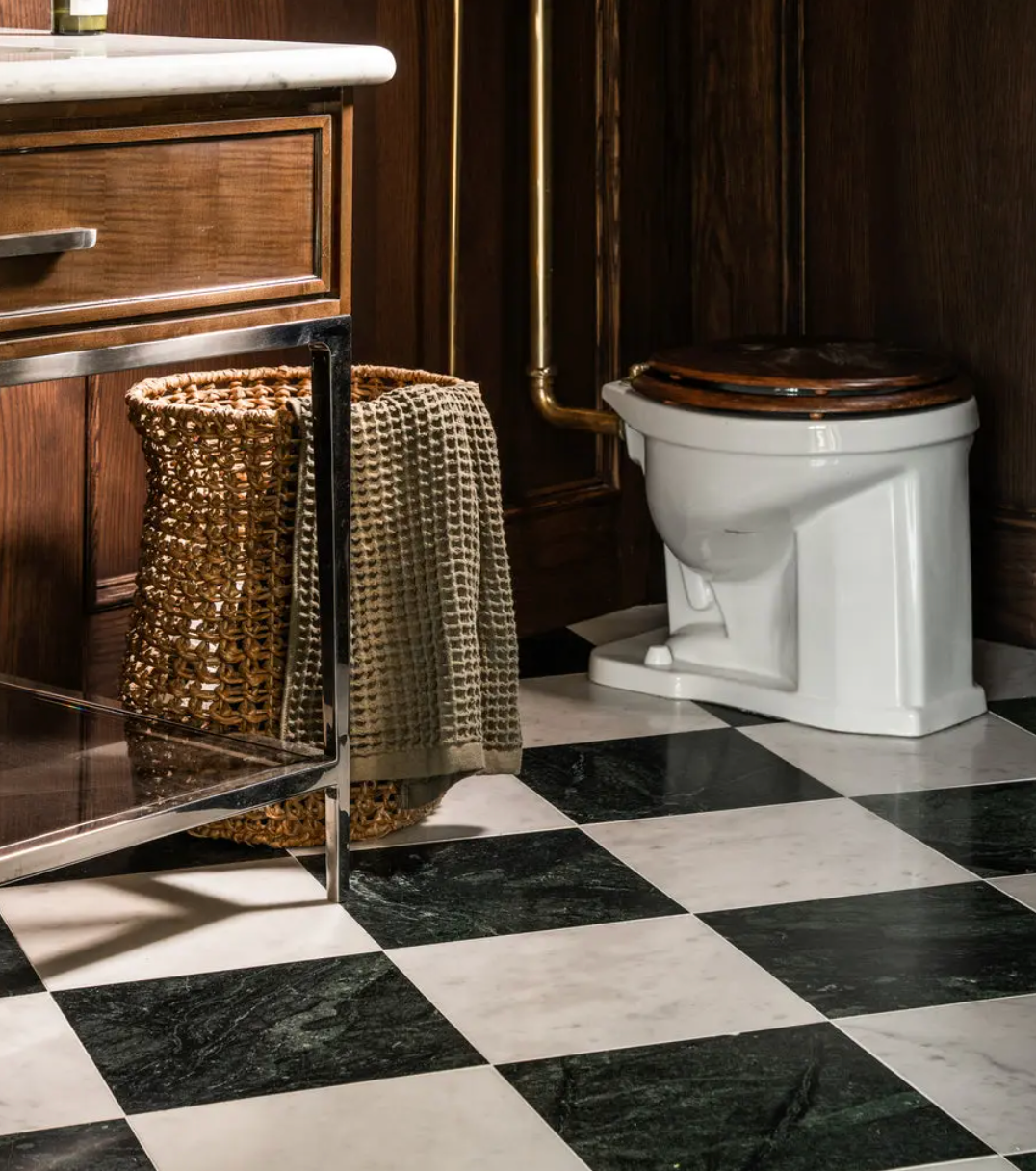 A bathroom with Verde Alpi 12x12 black and white handmade tile by Zia Tile, a white toilet with a dark wood seat, a woven basket, textured towel, and a metal-framed vanity topped with marble.