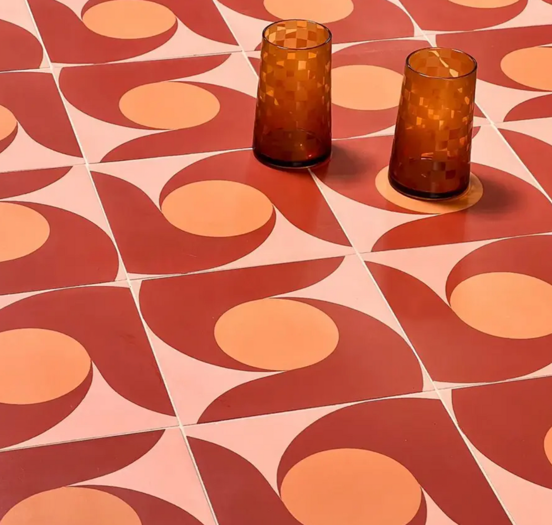 Two amber-colored glasses rest on a surface covered with Zia Tile's San Juan Delta Moon 8x8 tiles, featuring a retro-inspired geometric pattern in shades of red, orange, and pink with circular and curved shapes.