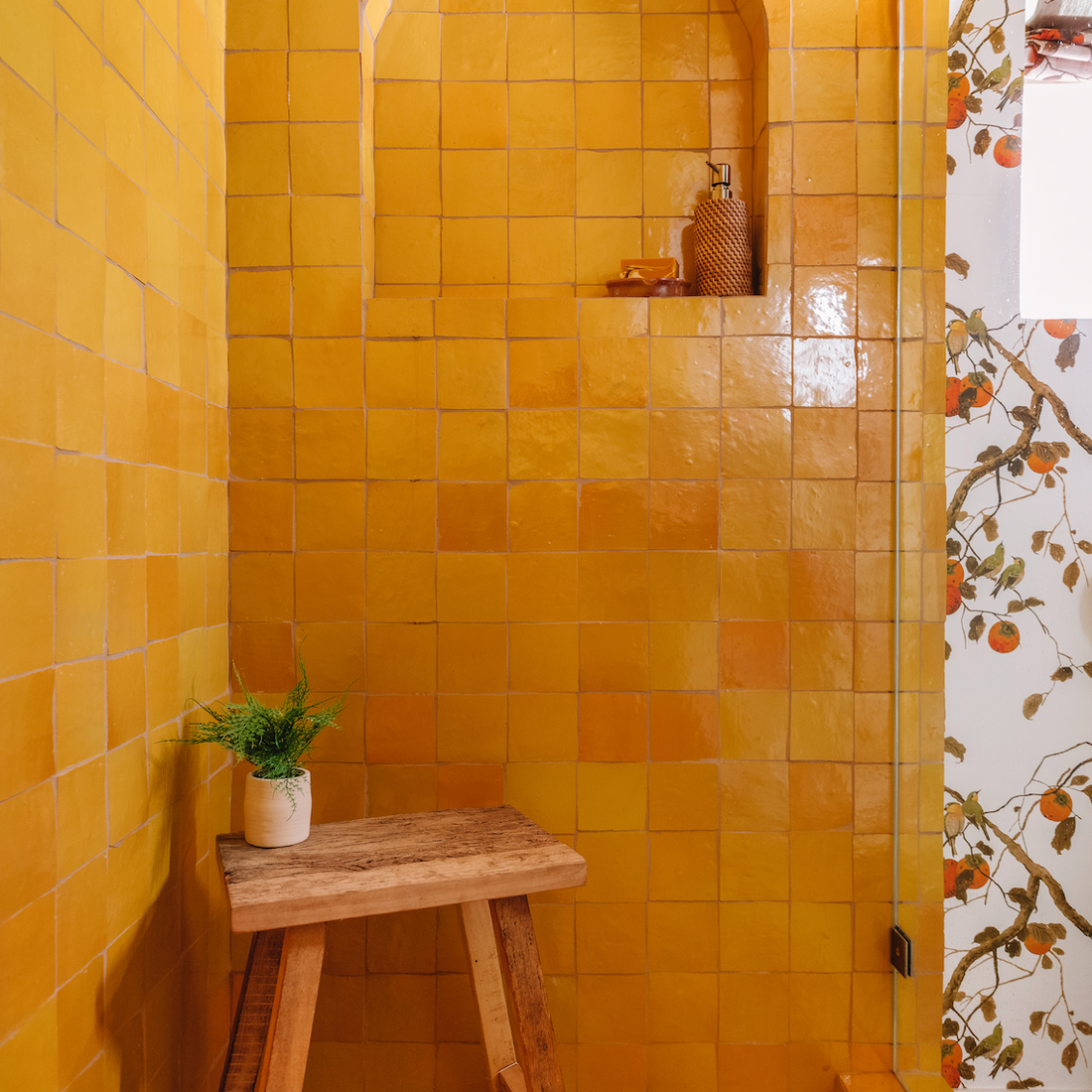 A shower area with glossy yellow square tiles, featuring an arched niche holding a decorative item, a wooden stool with a potted plant, and part of a floral wallpaper visible on the adjacent wall.