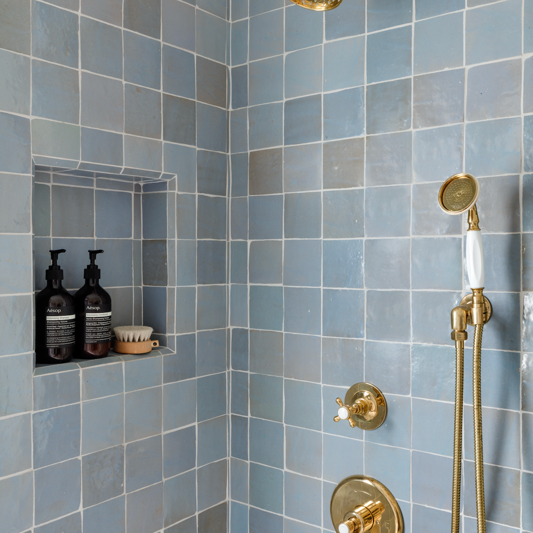 Shower with square blue tiles, gold showerhead and fixtures, a handheld shower, and a built-in wall niche holding two bottles and a soap dish.