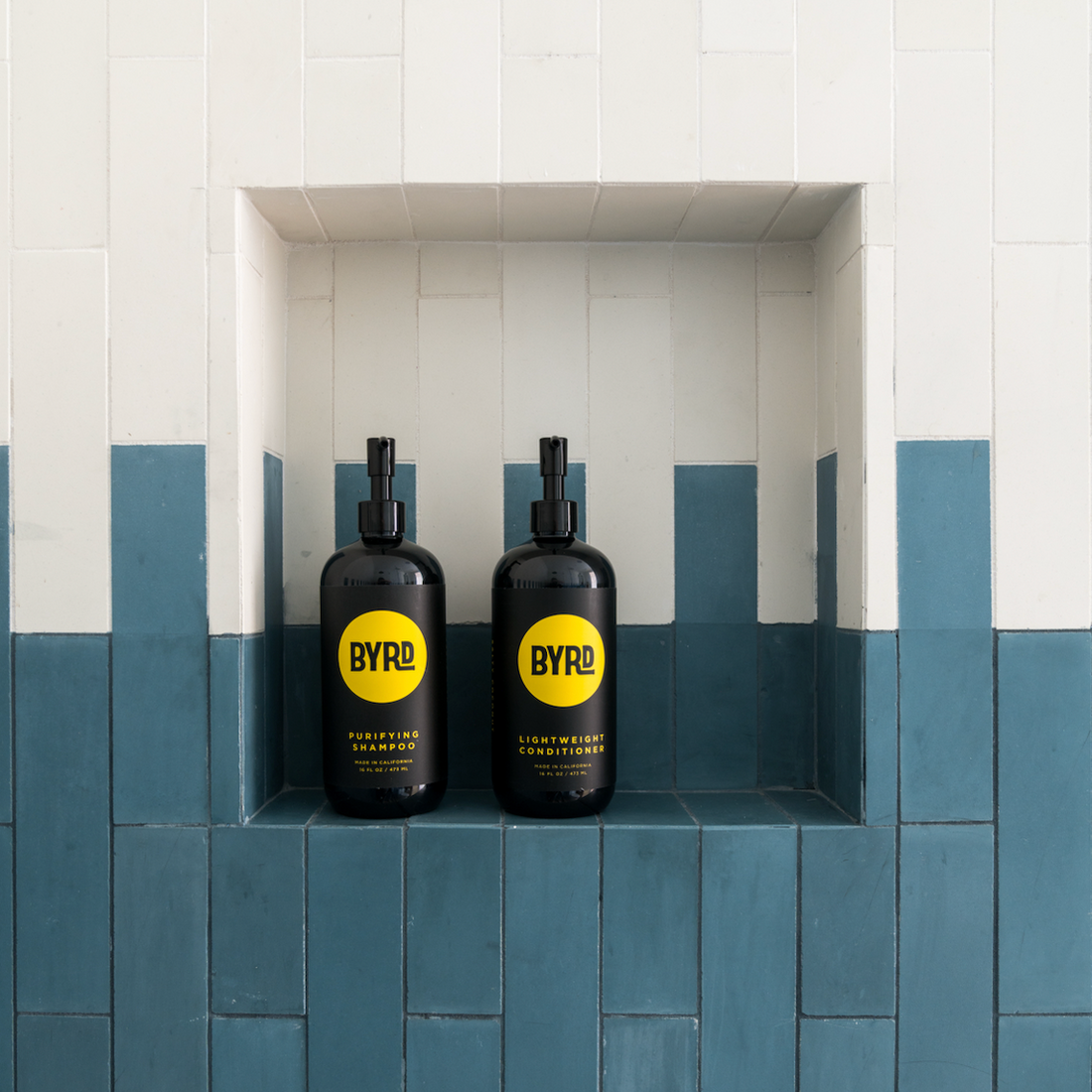 Two black BYRD shampoo and conditioner bottles with yellow labels are placed side by side in a recessed shelf of a tiled wall, featuring white and teal-blue rectangular tiles.