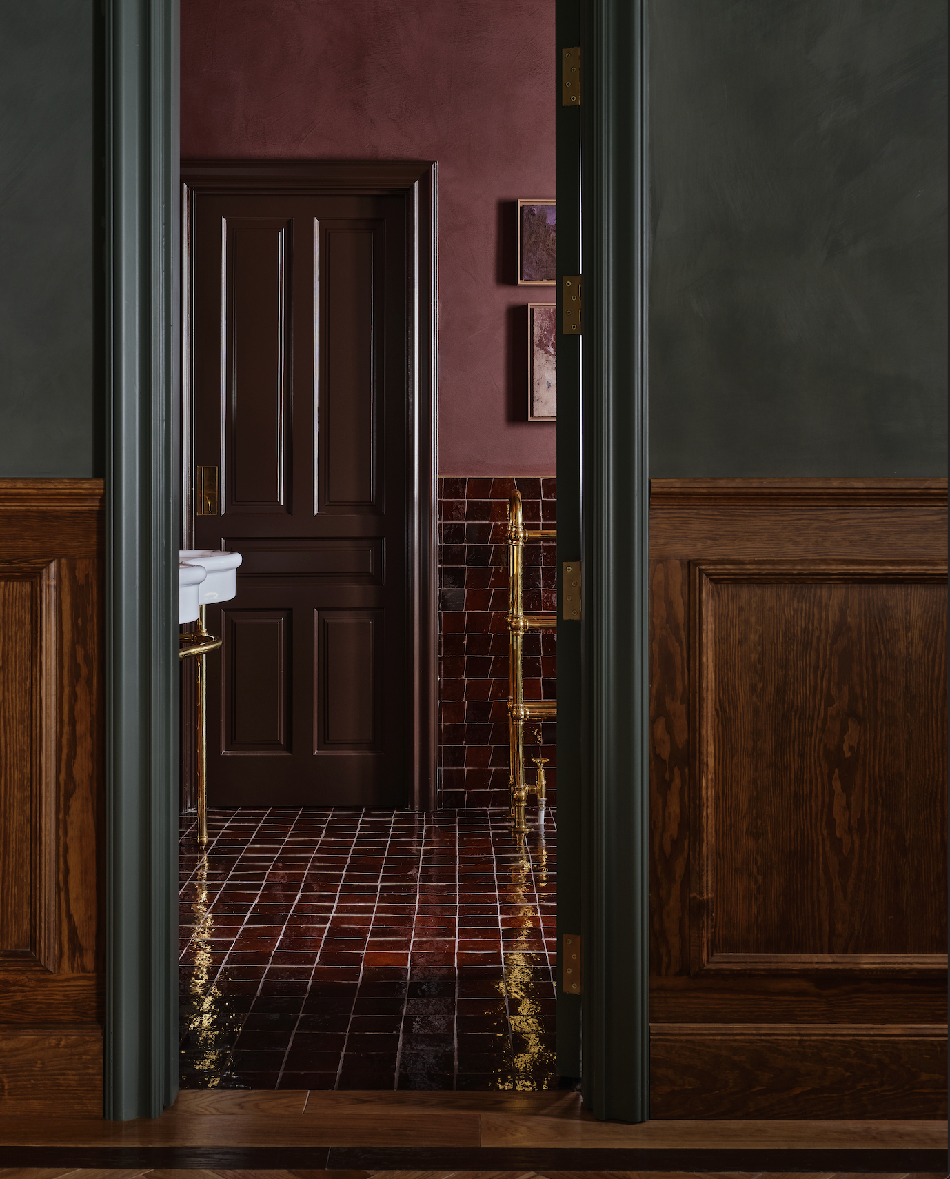 View through an open doorway into a bathroom with glossy burgundy tiles on the lower wall and floor, a dark brown wooden door, deep red painted upper walls, a white sink, and rich wooden wall paneling.