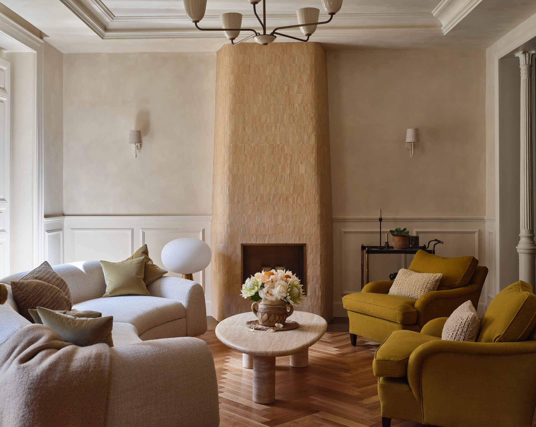 A cozy living room with a tan brick fireplace, cream sofas, mustard yellow armchairs, a round marble coffee table with a flower arrangement, wood flooring, and neutral walls with classic trim and molding.