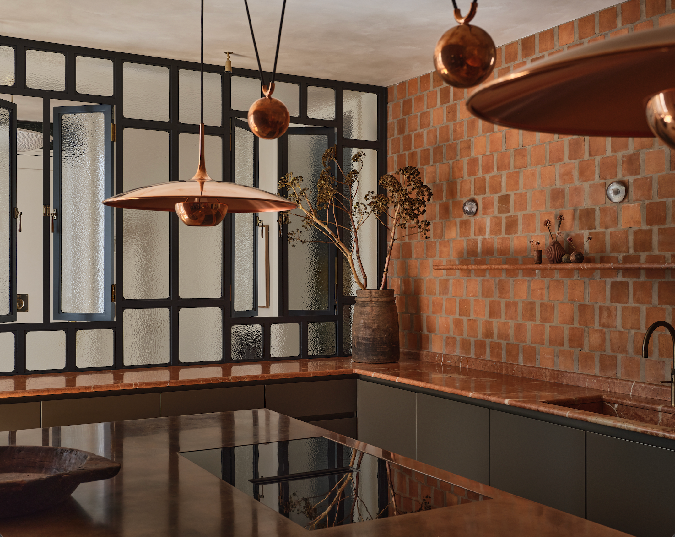 A modern kitchen with warm terracotta tiles, copper pendant lights, sleek dark cabinets, and large windows with frosted glass. A vase with dried branches sits on the counter, adding a rustic touch.