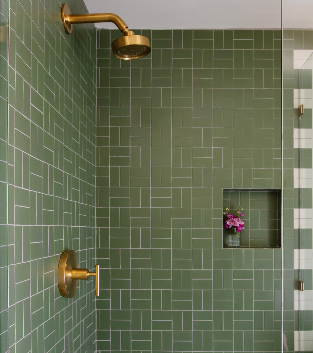 A shower features Sequoia 2x4 matte green geometric ceramic tiles by Zia Tile, paired with a gold showerhead and handle. A built-in shelf holds a vase with purple flowers.