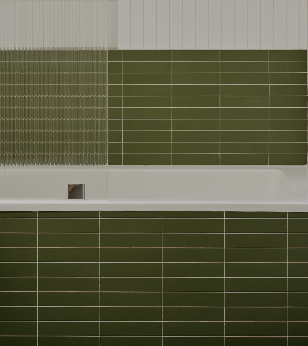A bathtub front and wall backsplash covered in olive green Sequoia 2x8 ceramic tiles by Zia Tile, next to a fluted glass shower screen.