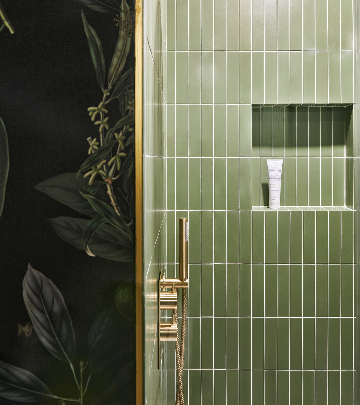 Sequoia 2x8 ceramic tiles by Zia Tile line a modern shower with a built-in shelf holding a white tube. Gold fixtures and a dark floral-patterned wall appear beside the matte finish tiles.