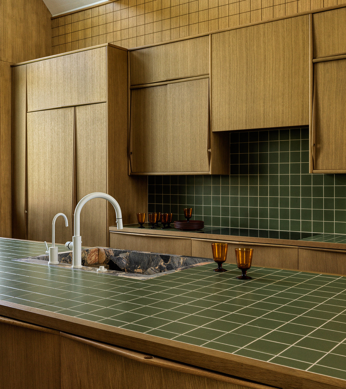 A modern kitchen features Zia Tile's Sequoia 4x4 green ceramics on countertops and backsplash, wooden cabinets, a white faucet, two amber glasses, and a black and brown stone sink in a warm, well-lit space.