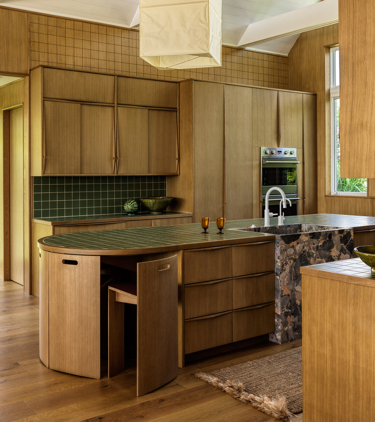 A modern kitchen with light wood cabinets, an island and backsplash tiled in Sequoia 4x4 by Zia Tile, built-in oven, large window, marble countertop, and minimalist decor featuring two amber glasses and a green bowl.