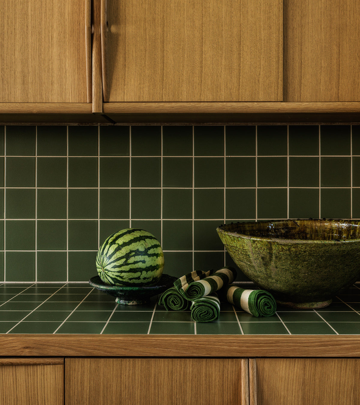 A kitchen counter features Sequoia 4x4 green ceramics by Zia Tile, a watermelon on a black plate, a green ceramic bowl, and several rolled-up green napkins. Wooden cabinets are above and below the counter.