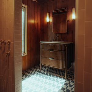 A cozy bathroom featuring Zia Tile’s Sidecar Black 4x8 tile floor, wood-paneled walls, a wooden vanity with mirror, wall-mounted lights, and natural light streaming through a window.