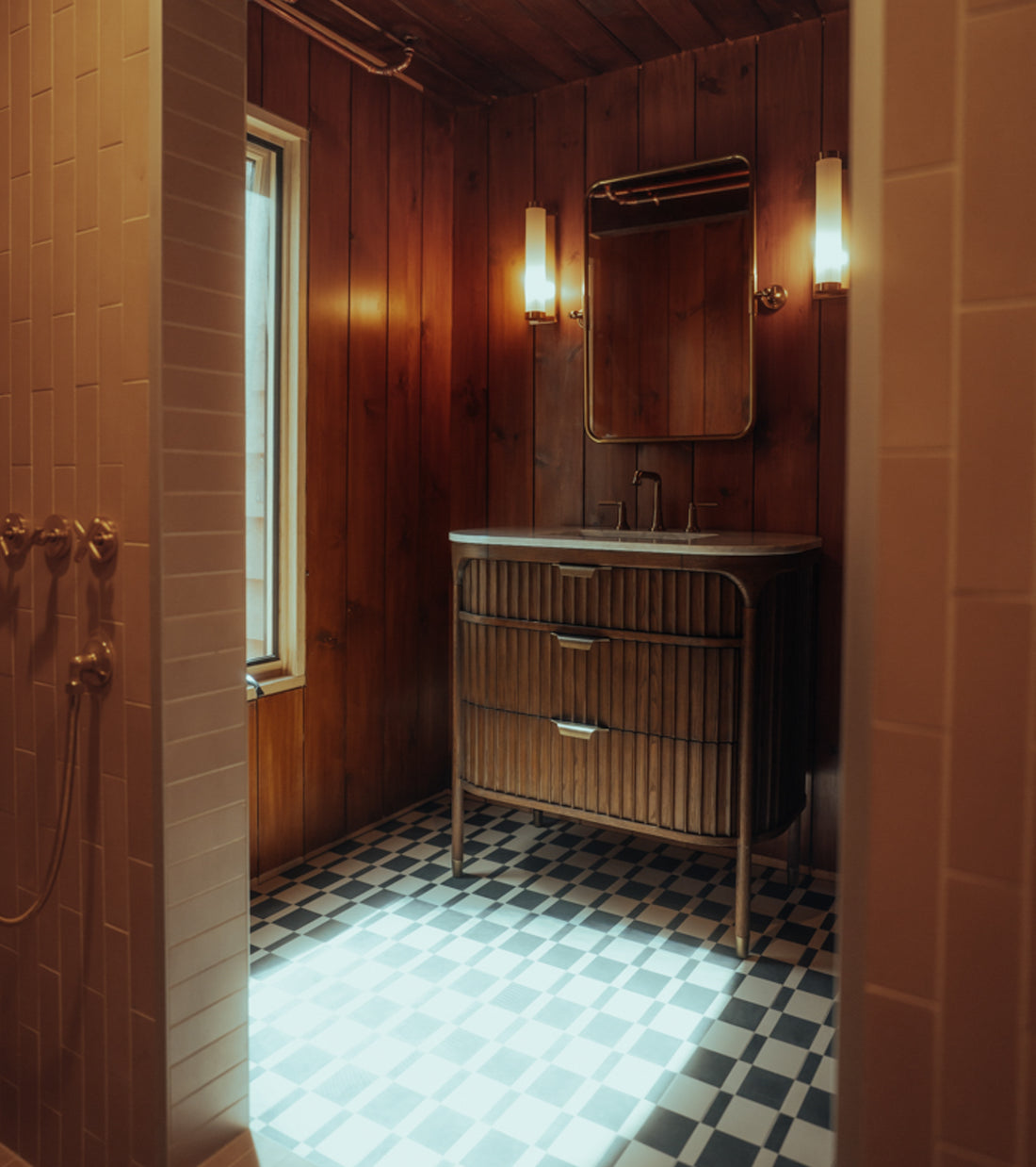 A cozy bathroom featuring Zia Tile’s Sidecar Black 4x8 tile floor, wood-paneled walls, a wooden vanity with mirror, wall-mounted lights, and natural light streaming through a window.