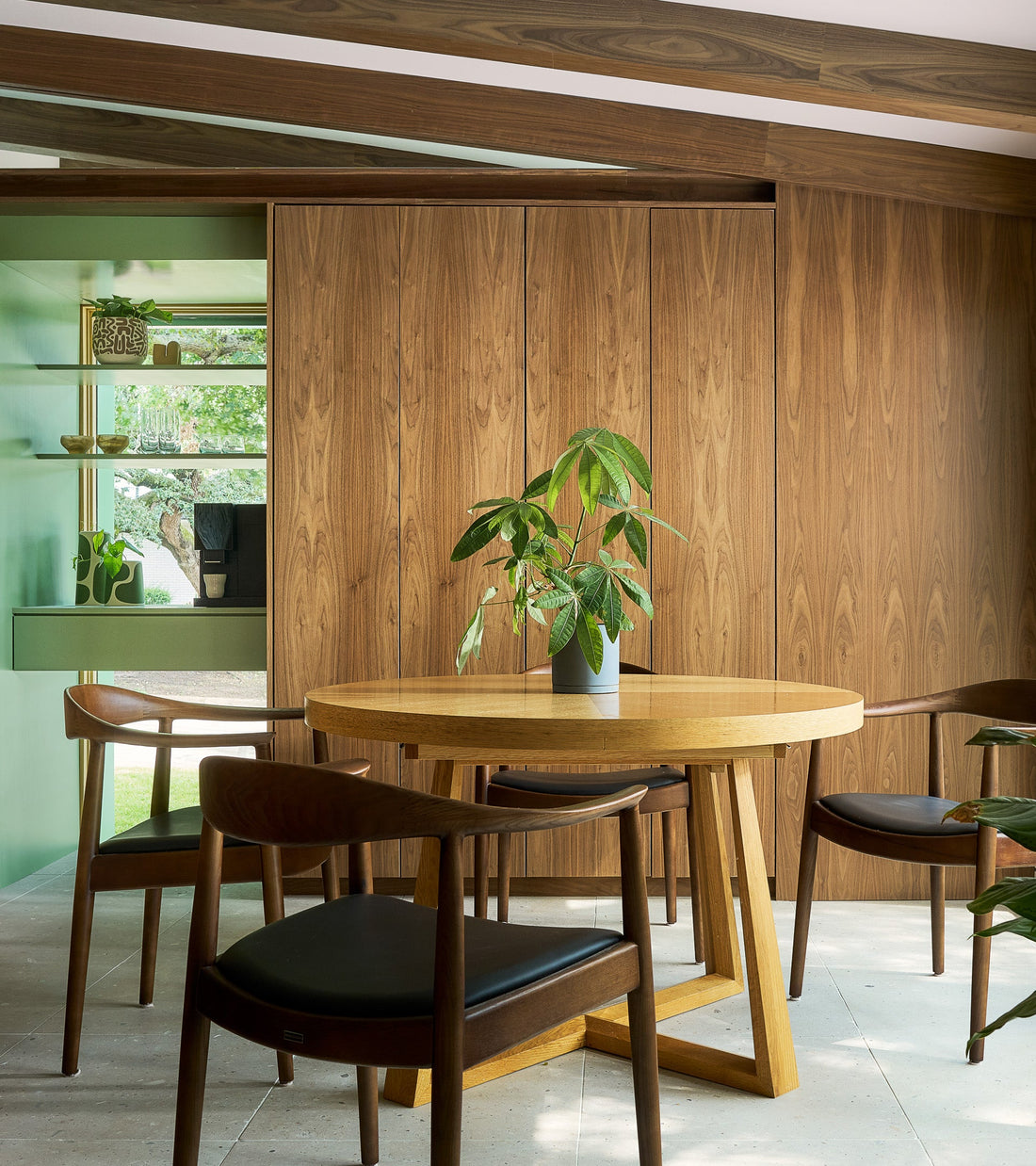 A modern dining area with a round wooden table, four dark-wood chairs with black seats, a potted plant on the table, wood-paneled walls, and shelves with decor items beside a window showing greenery outside.