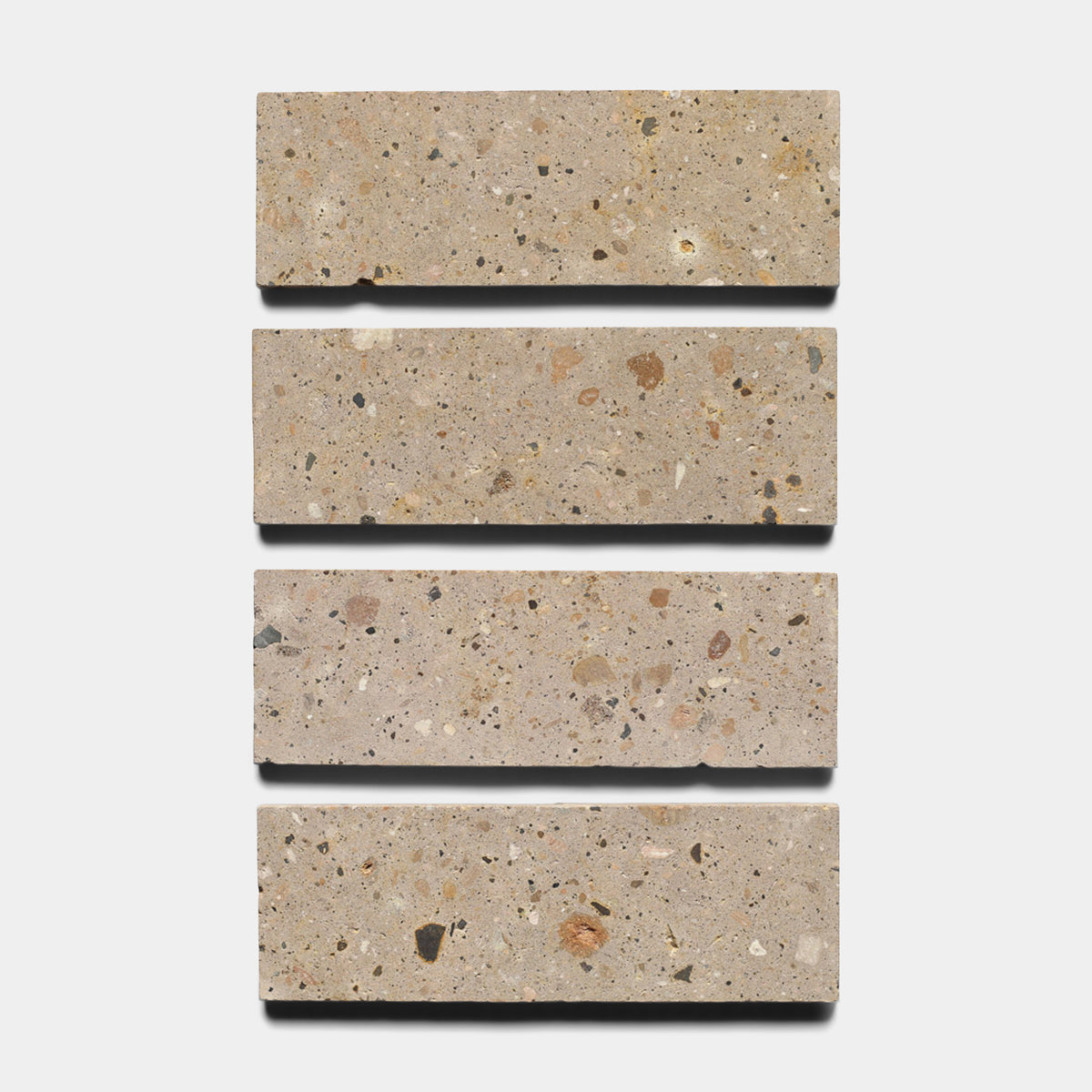 Four Sierra 4x12 terrazzo tiles by Zia Tile, featuring a light beige background with multicolored stone fragments, are arranged vertically and evenly spaced on a white backdrop, echoing the natural beauty of Mexican Cantera stone.