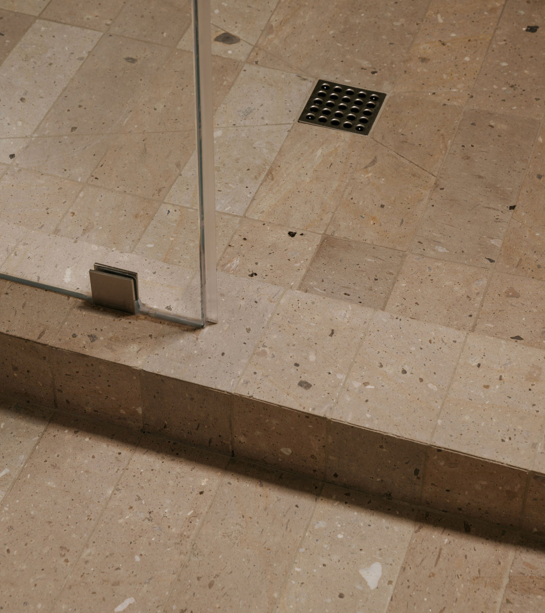 A shower floor and step covered in Sierra 4x12 rectangular, speckled beige Mexican Cantera stone tiles with a metal drain.