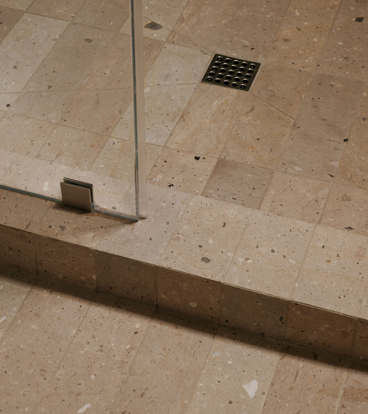 A shower floor and step covered in Sierra 4x12 rectangular, speckled beige Mexican Cantera stone tiles with a metal drain.