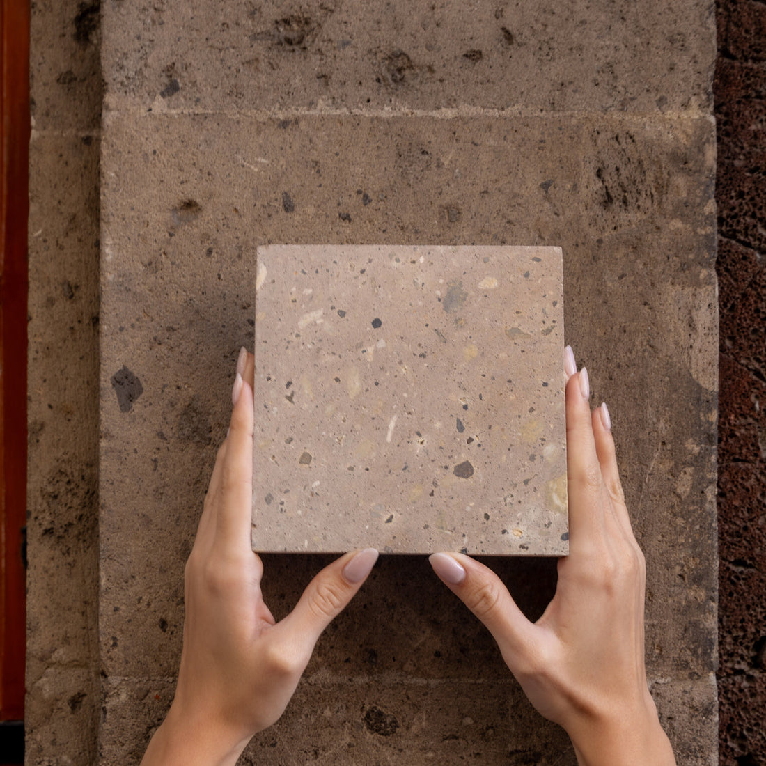 Two hands with manicured nails hold a square, light brown stone tile with small speckles, positioned against a larger, textured stone surface.