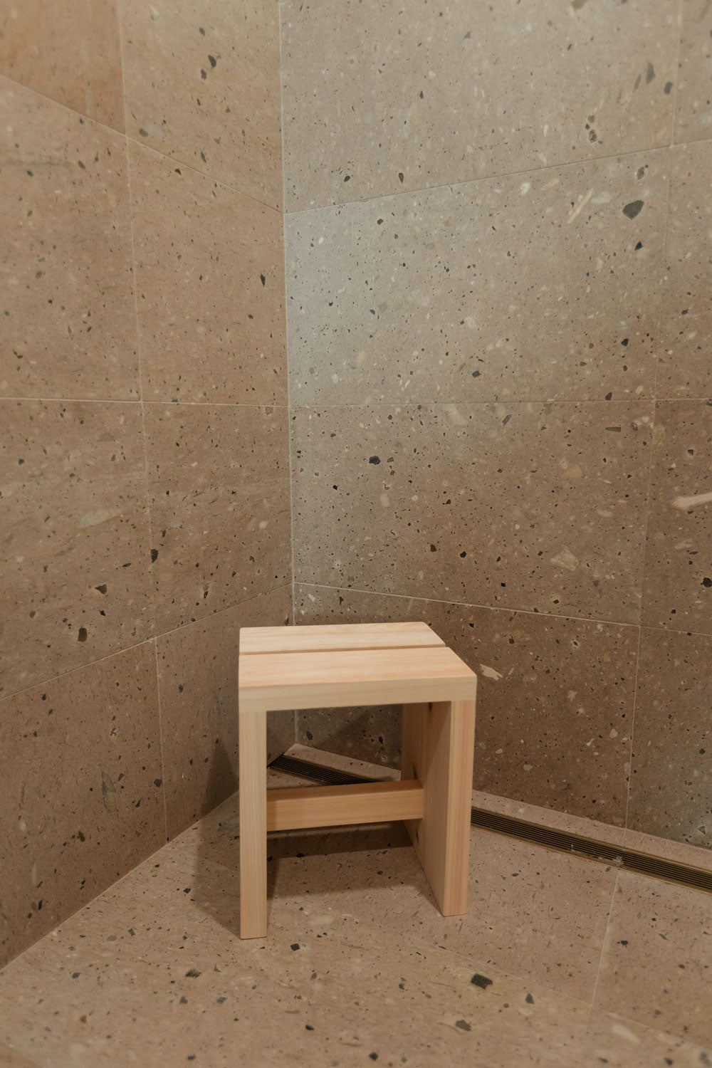 A small, light wooden stool is placed in the corner of a shower with beige, speckled stone walls and floors.