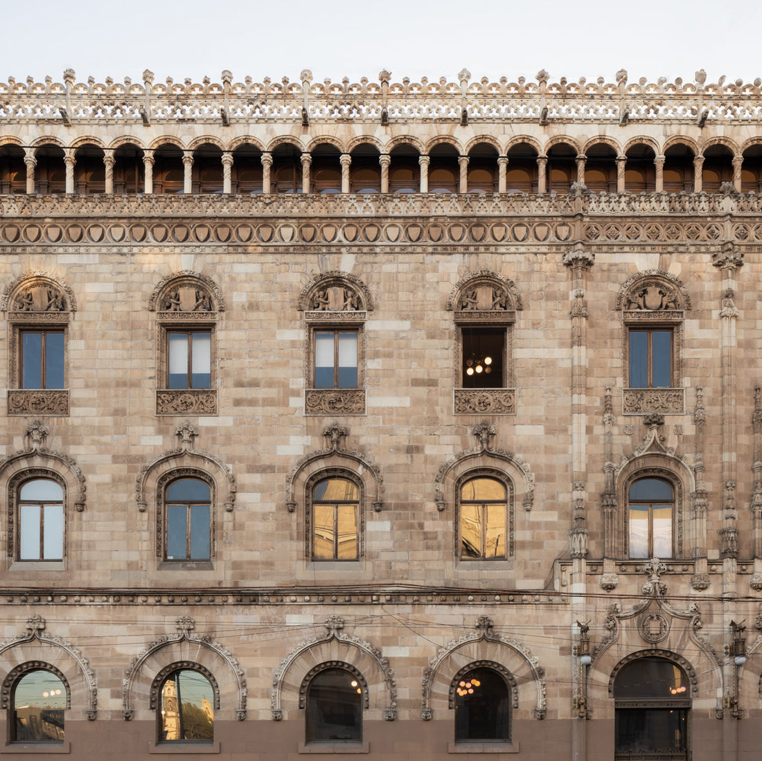 A historic stone building facade with arched and rectangular windows, intricate carvings, and detailed ornamentation along the roofline. Some windows reflect the sky, and warm lights are visible inside.