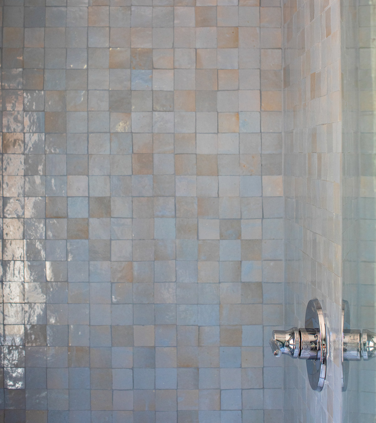 Close-up of a shower wall featuring glossy, light blue and beige Skylight 2x2 tiles by Zia Tile with a chrome shower control handle, highlighting the artisanal charm of handcrafted mosaic tiles.