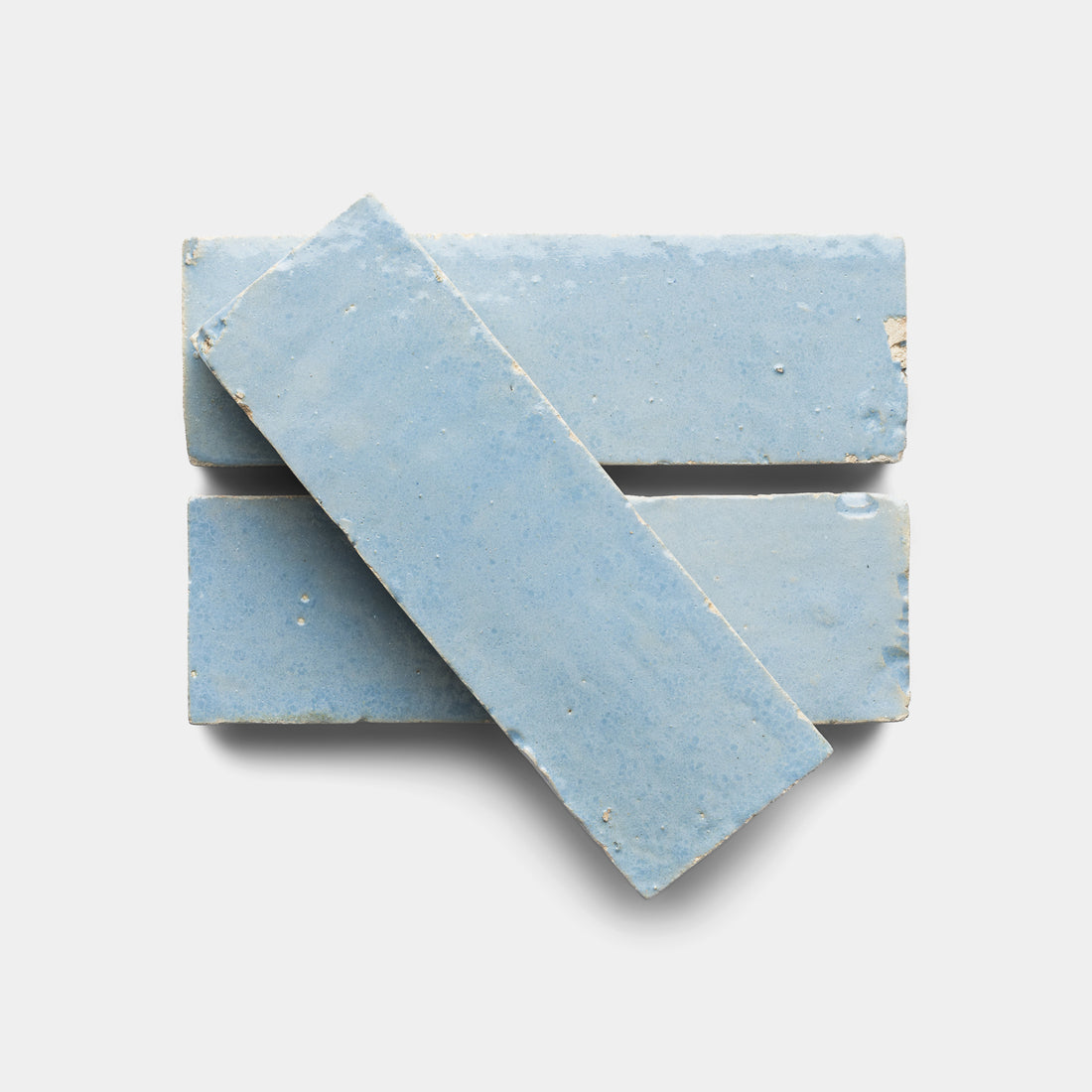 Three rectangular Skylight 2x6 zellige tiles from Zia Tile, in light blue, are stacked in a staggered arrangement on a white background. The top tile is angled diagonally across two horizontally placed tiles for a Moroccan-inspired look.
