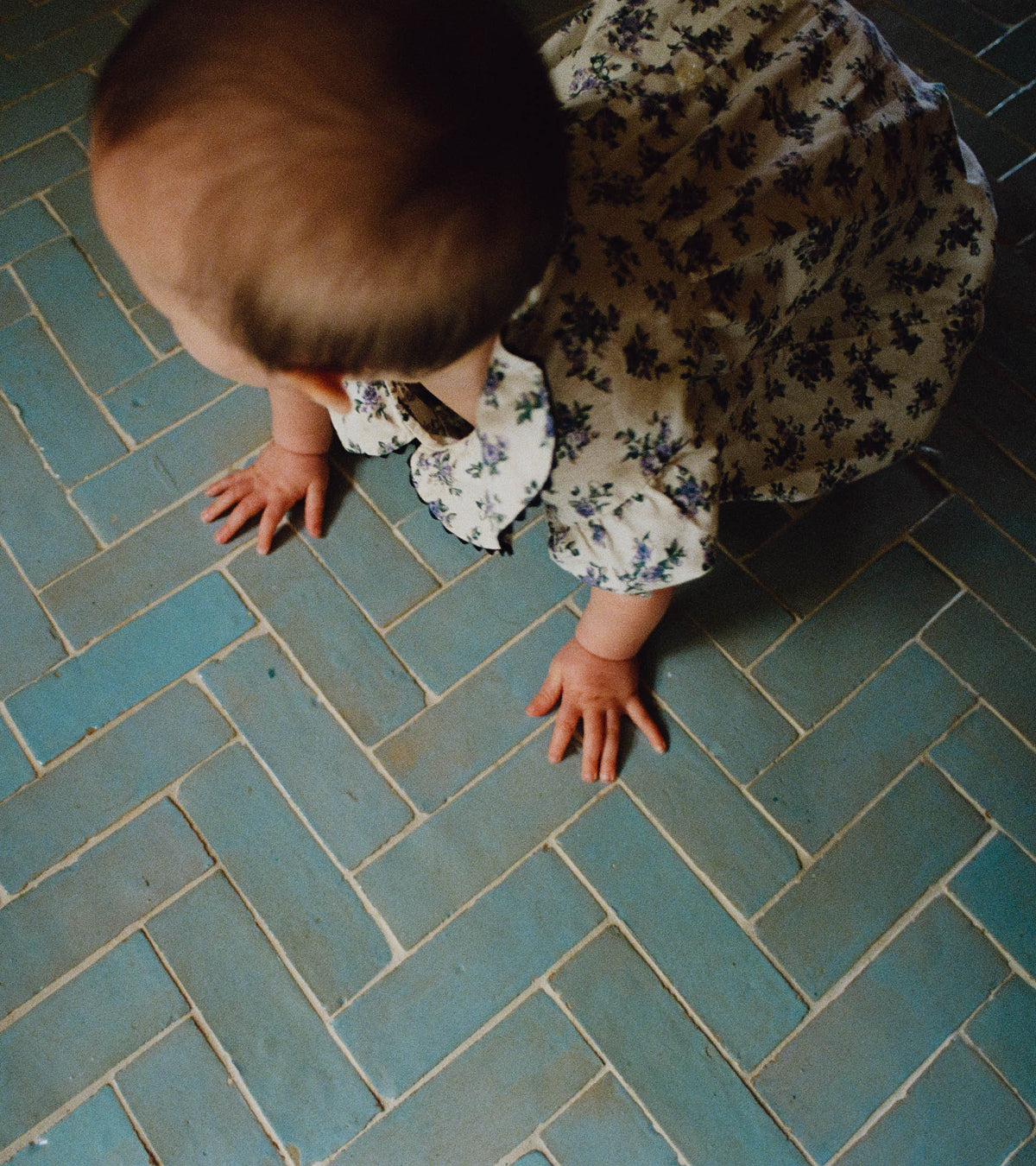 A baby in a floral dress crawls on a blue Zia Tile Skylight 2x6 floor, with only the top of the baby's head and hands visible.