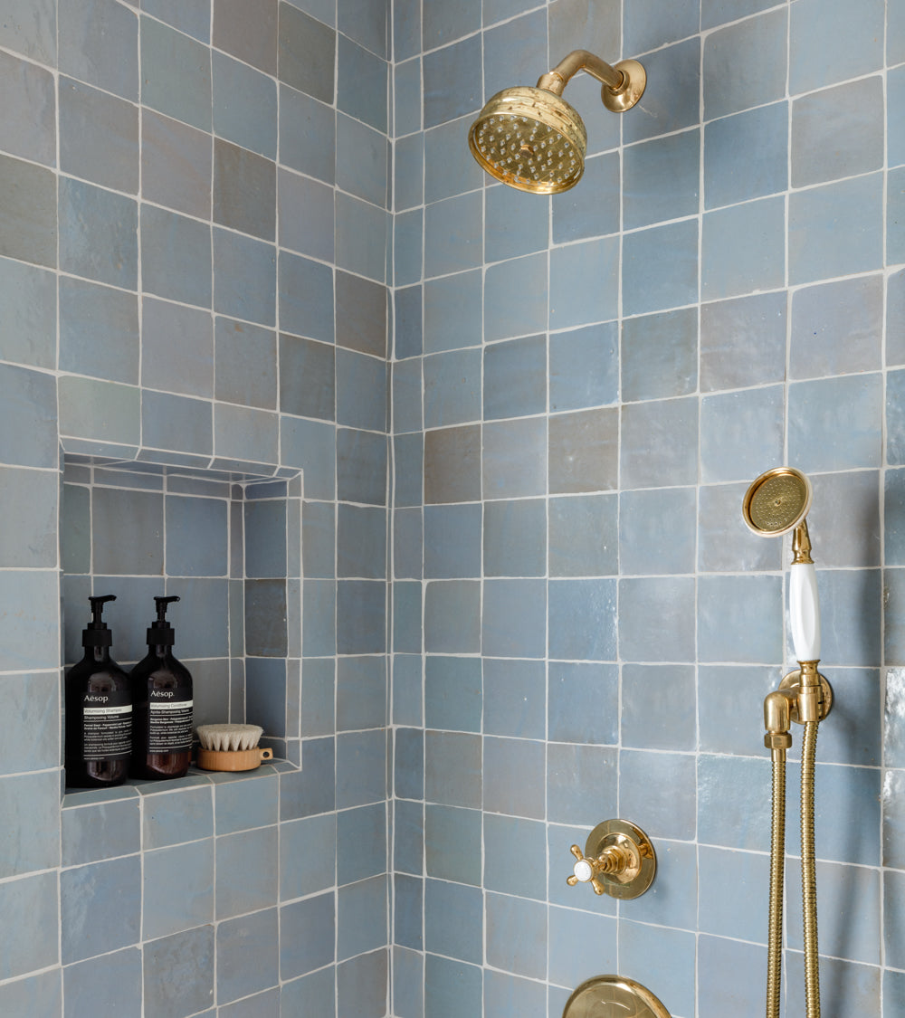 A shower featuring Skylight 4x4 tiles by vendor-unknown in glossy light blue, a gold showerhead, handheld sprayer, and a recessed shelf with two bottles and a soap dish evokes Moroccan tile style.