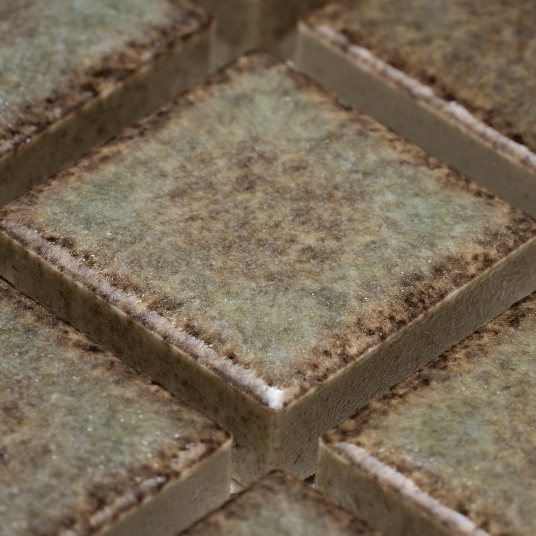 Close-up of Zia Tile’s Spanish Moss 1x1 tiles—square ceramic pieces with a mottled green, brown, and beige surface, slightly rough texture, and dark edges—arranged in a neat grid pattern.