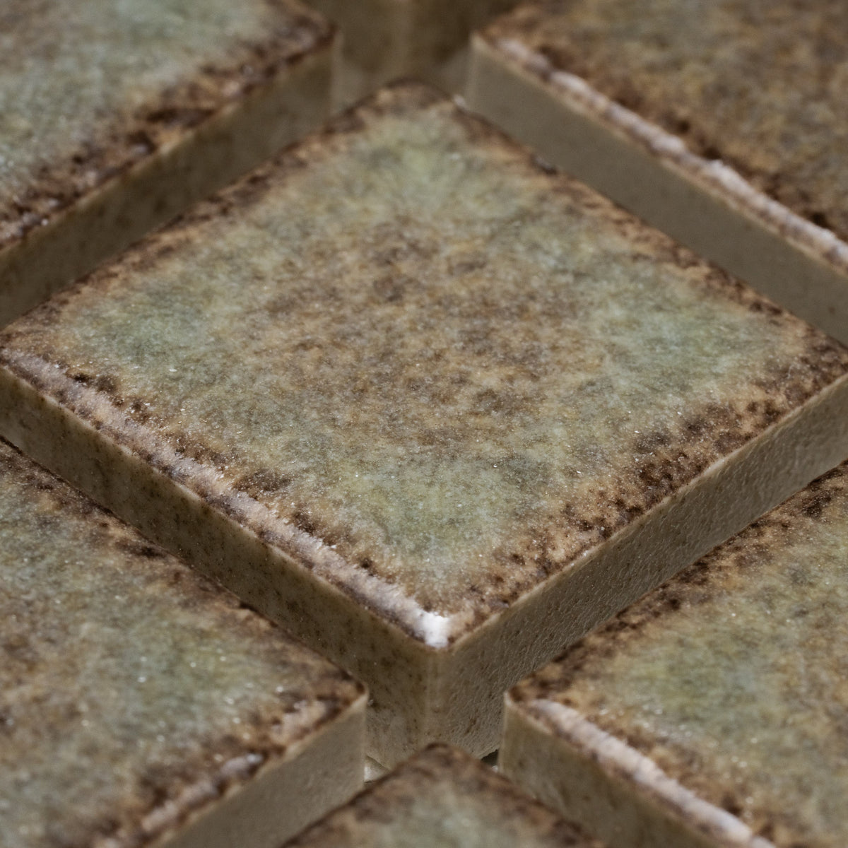 Close-up of Zia Tile’s Spanish Moss 1x1 tiles—square ceramic pieces with a mottled green, brown, and beige surface, slightly rough texture, and dark edges—arranged in a neat grid pattern.