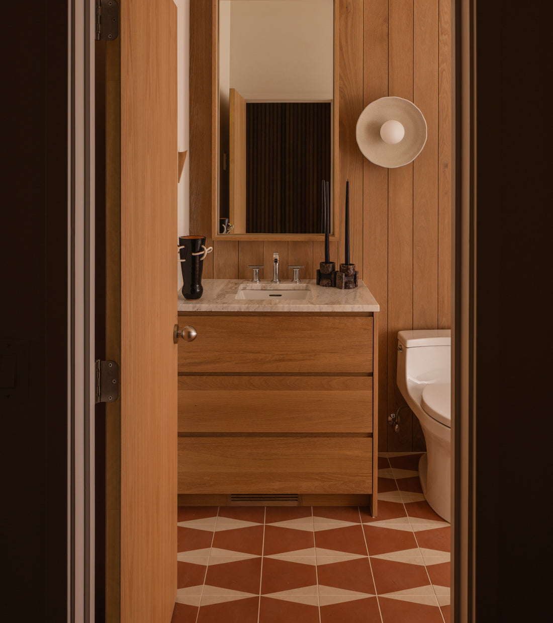 A modern bathroom features wood-paneled walls, a marble-topped wooden vanity, a round wall sconce, white toilet, large mirror above the sink, and artisan geometric flooring with Zia Tile's Sparrow Rust 8x8.