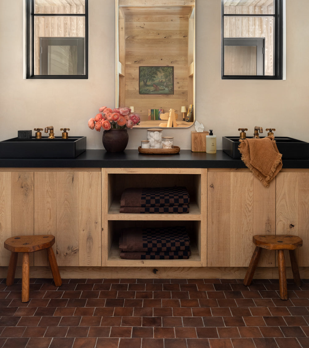 A modern bathroom featuring a double black sink vanity, light wood cabinets, two mirrors, brown checkered towels, pink flowers, soap, and wooden stools on 4x4 Square + Madera handmade Cotto terracotta floor tile by Zia Tile.