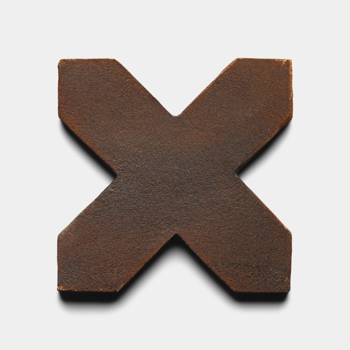 A large, textured, rusty metal letter X on a white background casts a faint shadow below, echoing the industrial charm of Zia Tile’s Stars & Cross + Madera sample.