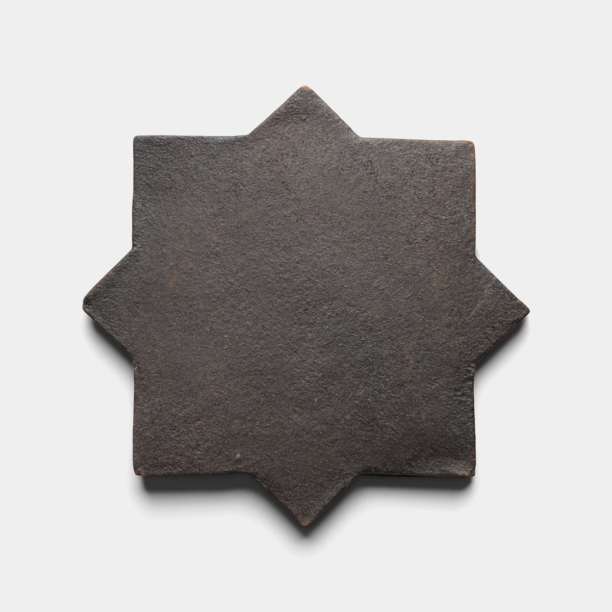 A dark, eight-pointed star-shaped Stars & Cross + Oscura tile by Zia Tile, featuring a rough, textured surface, is centered against a plain white background.