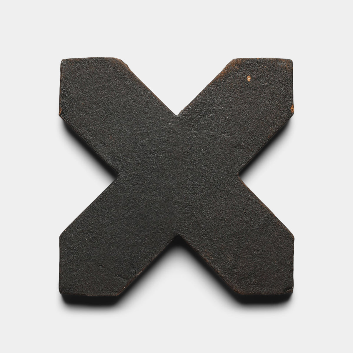 A Stars & Cross + Oscura sample tile from Zia Tile, with a black X-shaped design and textured cement finish, rests on a white background, casting a subtle shadow underneath.