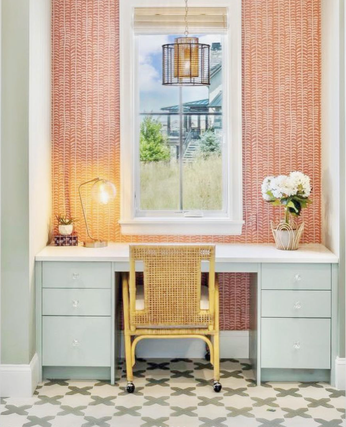 A stylish home office features a rattan chair at a pale blue desk with six drawers, a lamp, a vase of white flowers, patterned red wallpaper, and a window showing a grassy outdoor scene. Patterned tile floor adds contrast.