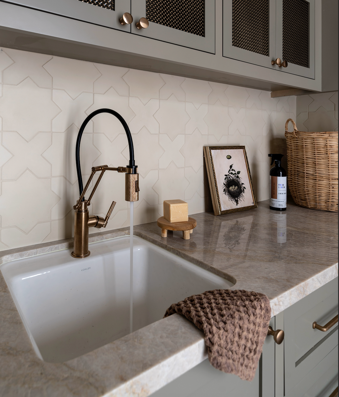 A modern kitchen sink features a bronze faucet, running water, beige soap on a wooden dish, framed art, soap bottle, wicker basket, and brown cloth on the counter. The Stars & Cross Bone cream tiles from vendor-unknown add elegance to the space.