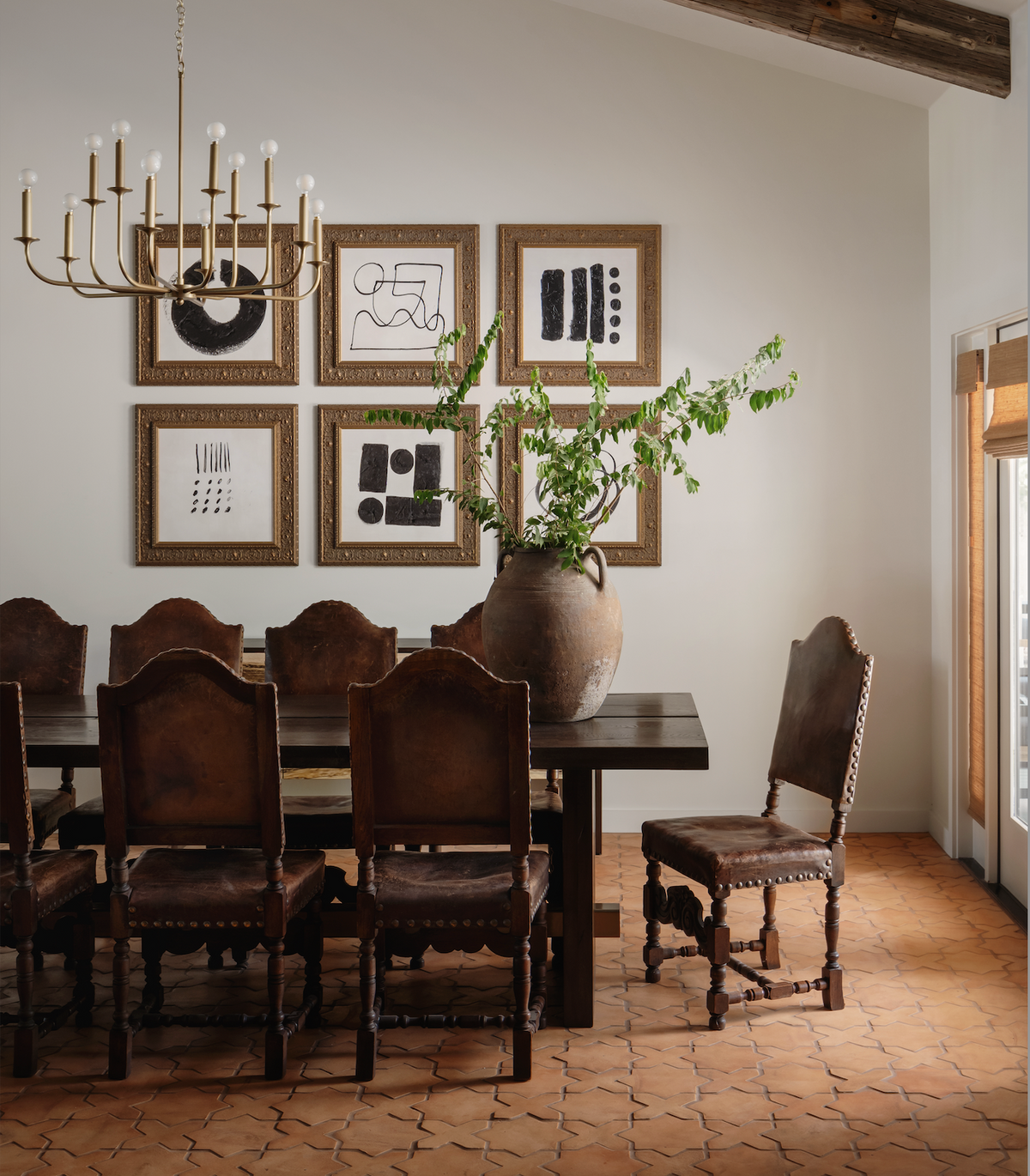 A dining room features a dark wood table with eight ornate chairs, a green-branch centerpiece, abstract black-and-white art, a gold chandelier, and Zia Tile’s Stars & Cross + Madera handmade tile flooring.