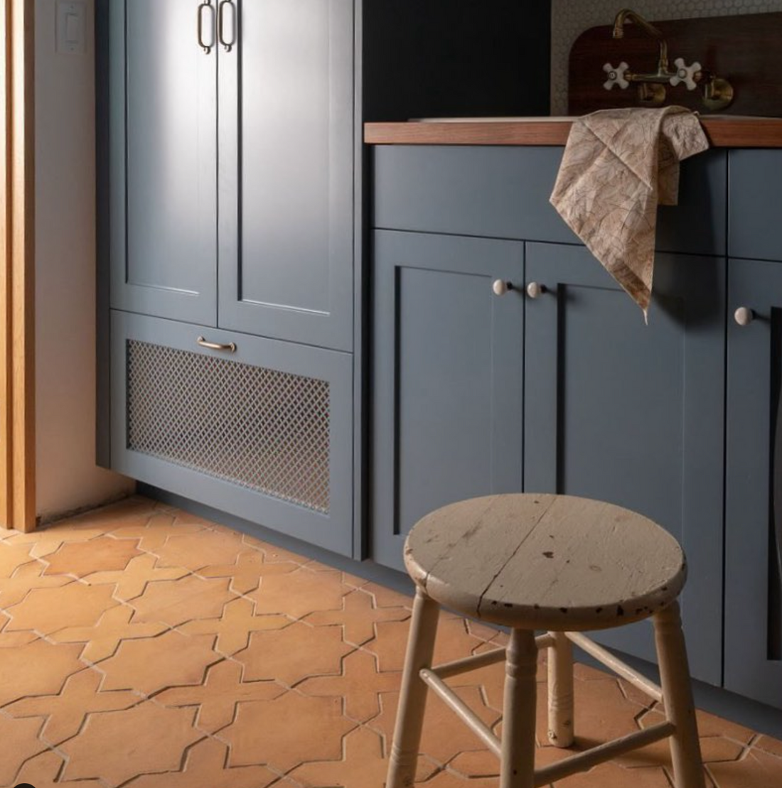 A rustic wooden stool rests on Zia Tile's Stars & Cross + Madera cement tiles beside blue cabinets and a wooden countertop with a beige towel draped over the side, softly lit from the left.